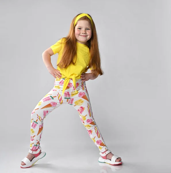 Studio shot van een vrolijk roodharig meisje in een zomer modieuze outfit, doe haar handen op haar riem en spreid haar benen wijd uit elkaar. Fijne vakantie momenten en oprechte kinderlijke emoties — Stockfoto