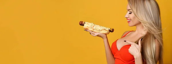 Λαμπερό σέξι ξανθιά κοιτάζει σαγηνευτικά σε ένα hot dog εκμετάλλευση στο χέρι της. — Φωτογραφία Αρχείου