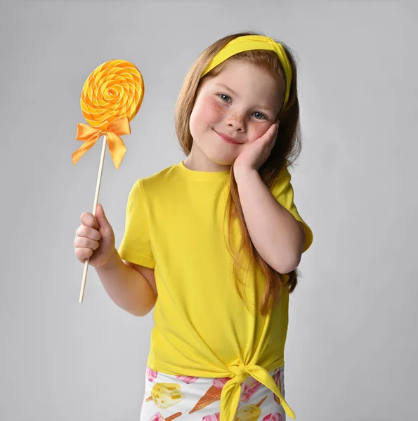 Roodharig schatje in een gele blouse en een hoofdband, is ontroerd en blij met haar koppelaarster - ze kreeg een grote heerlijke snoep. — Stockfoto