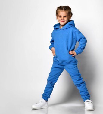Moda mavisi eşofmanlı küçük kız beyaz arka planda eğleniyor. Duygusal bir ifadeye sahip bir çocuk ellerini beline koyar. Çocuk sporları modasının tanıtımı.
