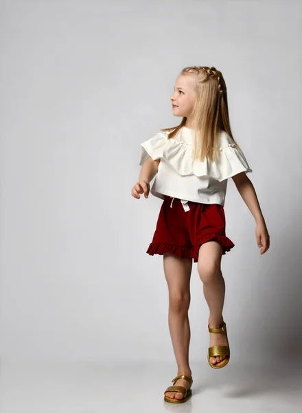 Χαριτωμένο χαμογελαστό κοριτσάκι σε μια λευκή μπλούζα με φούντες, μπορντό σορτς και σανδάλια, περπατά ζωηρά και κοιτάζει στο πλάι. — Φωτογραφία Αρχείου