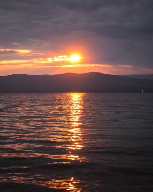 Denizde gün batımının fotoğrafı. Suda turuncu yansıma, arka planda dağlar. Dramatik manzara, nefes kesici deniz manzarası. Altın güneş ışınları.