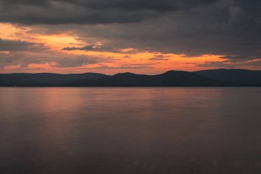 Denizde gün batımının fotoğrafı. Suda turuncu yansıma, arka planda dağlar. Dramatik manzara, nefes kesici deniz manzarası. Altın güneş ışınları.