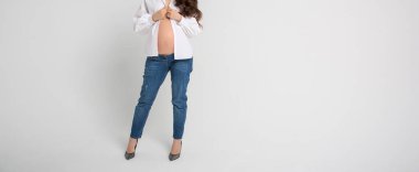 Uzun saçlı hamile bir kadın kot pantolon giyiyor, düğmeleri açık beyaz bir bluz ve beyaz arka planda yüksek topuklu ayakkabılar giyiyor.