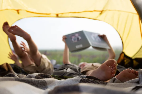 Children in a tent read a book lying down-a trip to nature in summer. In the frame of heels, legs and a book