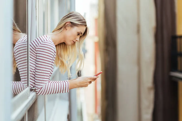 Attractive woman leaning out of her house window using a smartphone. — Stock Photo, Image