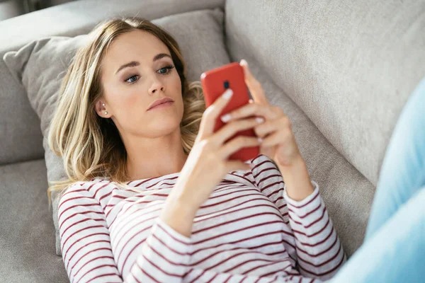 Mujer rubia joven usando su teléfono inteligente tumbado en el sofá. —  Fotos de Stock