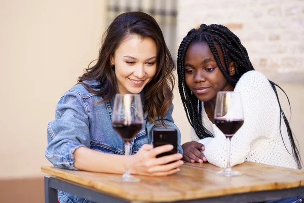 Twee vrouwen die samen naar hun smartphone kijken terwijl ze een glas wijn drinken. — Stockfoto