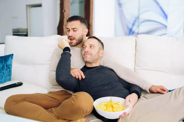 Couple gay assis sur le canapé à la maison regarder quelque chose à la télévision et avoir une collation. — Photo