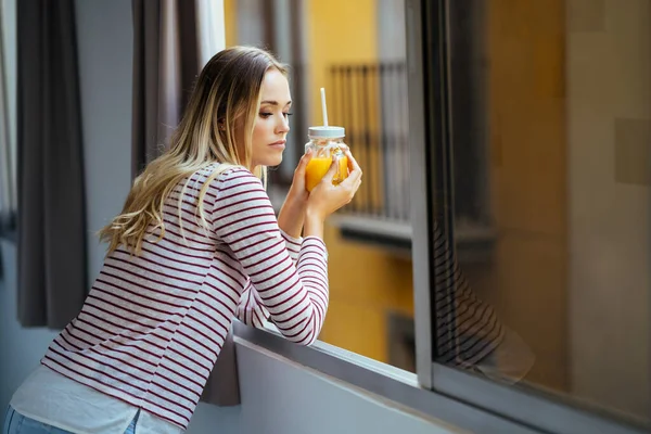 Mladá žena pije sklenici přírodní pomerančové šťávy, naklání se z okna svého domu. — Stock fotografie