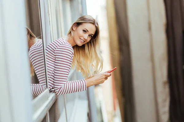 Jovem mulher se inclinando para fora de sua janela de casa usando um smartphone. — Fotografia de Stock
