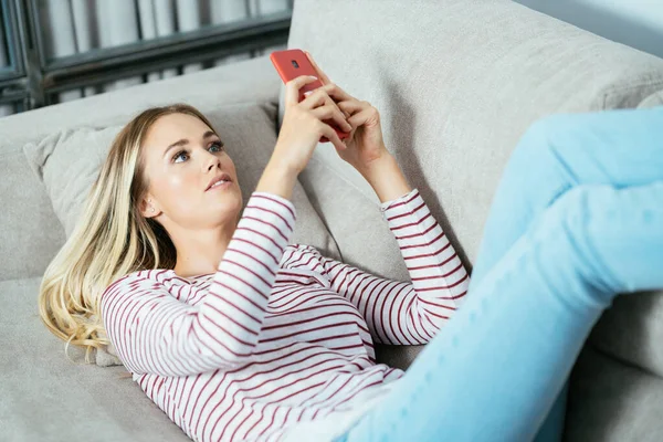 Молода біла жінка, використовуючи свій смартфон, лежить на дивані . — стокове фото