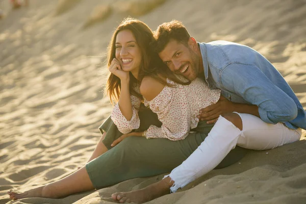 Pareja joven riendo juntos sentados en la arena de la playa — Foto de Stock