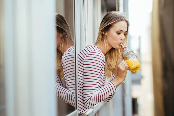 Junge Frau trinkt ein Glas natürlichen Orangensaft und lehnt sich aus dem Fenster ihres Hauses. — Stockfoto