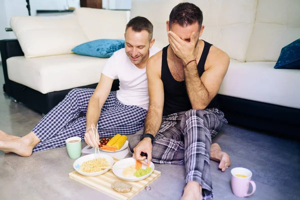 Gay paar eten samen zitten op hun woonkamer vloer. — Stockfoto