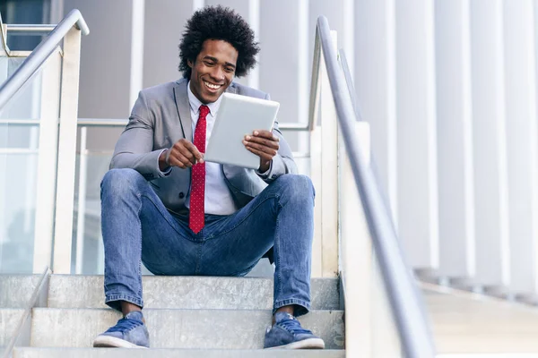 Hombre de negocios negro usando una tableta digital sentada cerca de un edificio de oficinas. —  Fotos de Stock