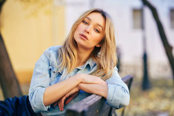 Mulher loira vestindo camisa jeans e saia de couro preto sentado em um banco urbano. — Fotografia de Stock