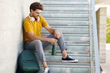 Erkek dışarıda alüminyum su şişesi, kulaklık ve sırt çantası kullanarak oturuyor..