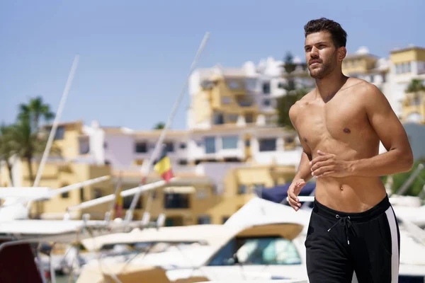 Młody człowiek ze sportowym ciałem biegnący bez koszulki przez port. — Zdjęcie stockowe