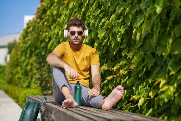 Masculino sentado fora usando uma garrafa de água de alumínio, fones de ouvido e mochila. — Fotografia de Stock