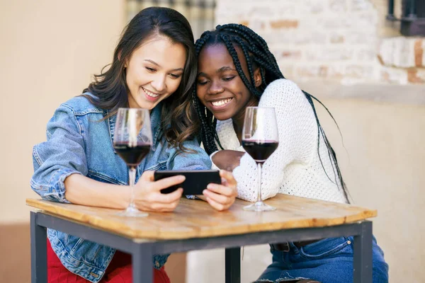 Twee vrouwen die samen naar hun smartphone kijken terwijl ze een glas wijn drinken. — Stockfoto