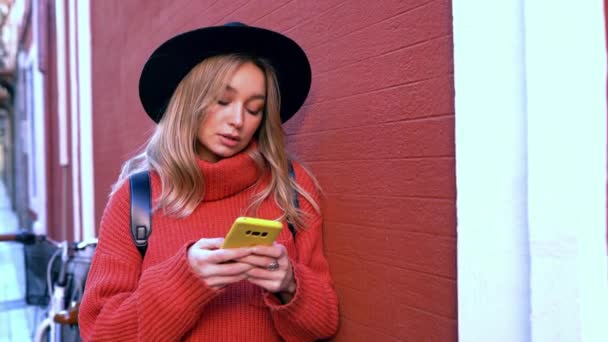 Mujer rubia escribiendo y leyendo mensajes de texto en su teléfono inteligente frente a una pared roja. — Vídeos de Stock