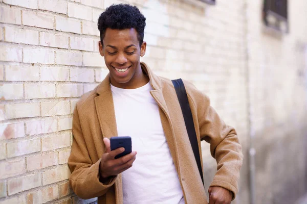 Giovane uomo nero digitando sul suo smartphone appoggiato a un muro di mattoni in strada. — Foto Stock