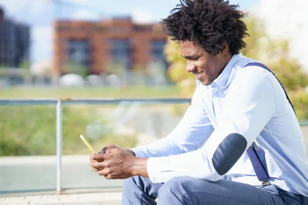 Uomo nero con acconciatura afro utilizzando uno smartphone seduto vicino a un edificio per uffici. — Foto Stock