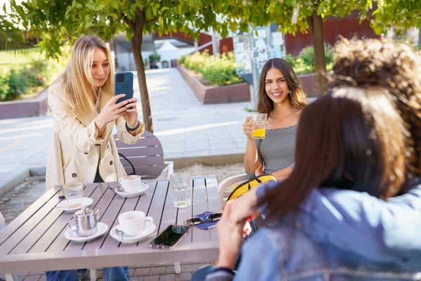 Chica rubia joven tomando una foto con un teléfono inteligente de sus amigos sentados en una mesa de bar. — Foto de Stock