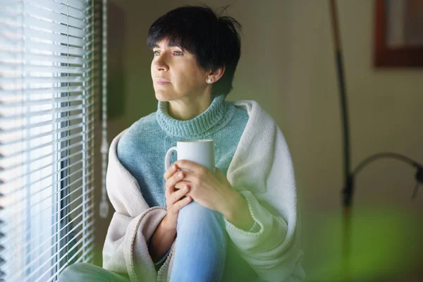 Mujer adulta tomando café y mirando por la ventana en el día de invierno. — Foto de Stock