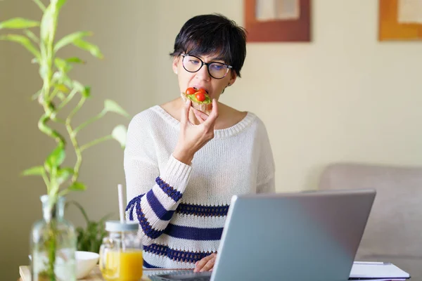Mulher comendo alguma comida saudável, enquanto se teletrabalho de casa em seu laptop. — Fotografia de Stock