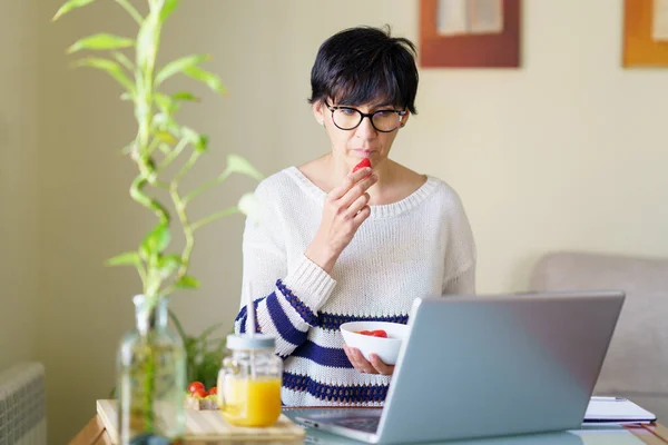 Mulher comendo morangos enquanto se teletrabalho de casa em seu laptop — Fotografia de Stock