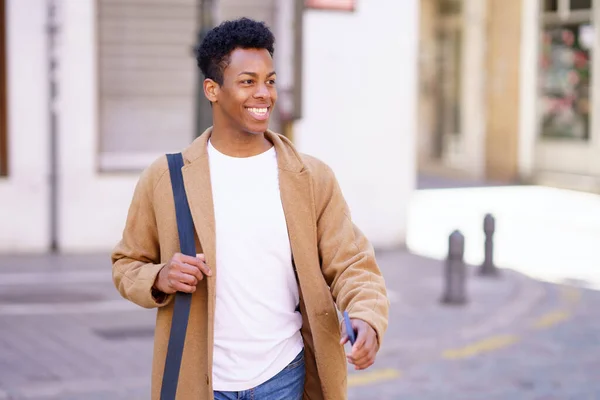 Felice uomo nero che cammina per strada portando in mano una valigetta e uno smartphone. — Foto Stock