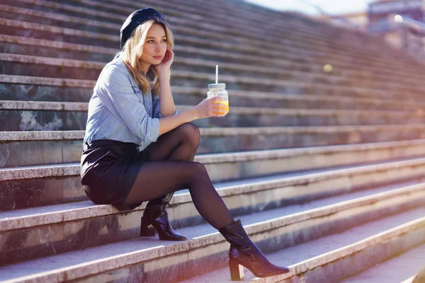 Mujer rubia con boina, bebiendo un zumo de naranja natural en un vaso de cristal, sentada en unos escalones. — Foto de Stock