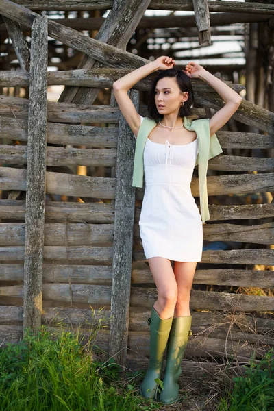 Azjatka, pozująca w pobliżu suszarni tytoniu, ubrana w białą sukienkę i zielone wellie. — Zdjęcie stockowe
