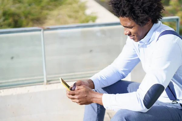 Black man with afro hairstyle using a smartphone sitting near an office building. — Stock Photo, Image