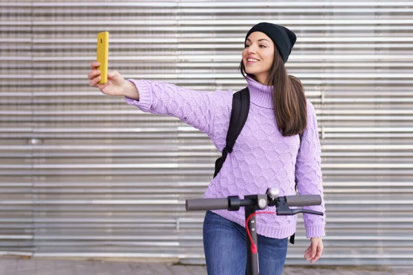 Mulher de vinte anos com scooter elétrico tirando uma selfie com um smartphone ao ar livre. — Fotografia de Stock