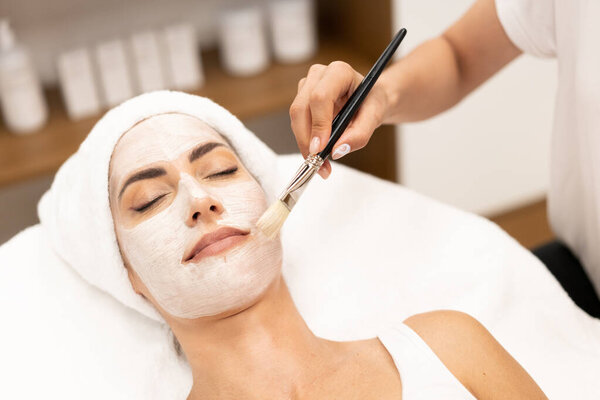 Aesthetics applying a mask to the face of a Middle-aged woman in modern wellness center.