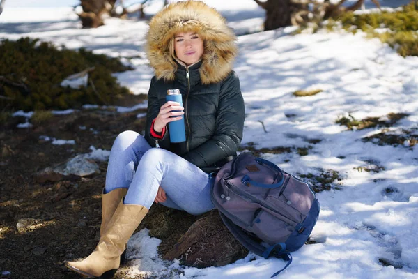 Frau trinkt etwas Heißes aus einer metallenen Thermosflasche und sitzt auf einem Felsen in den schneebedeckten Bergen. — Stockfoto