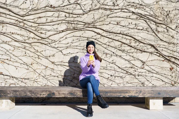 Mujer joven feliz sonriendo sentado en un banco al aire libre usando un teléfono inteligente. —  Fotos de Stock