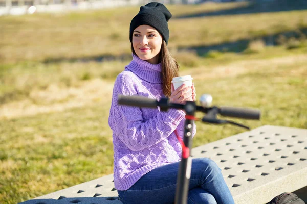 Female student with electric scooter taking a coffee break sitting on a bench outside her college. — Stock Photo, Image