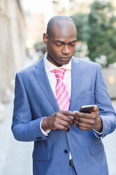Uomo d'affari nero che legge il suo smartphone in background urbano — Foto Stock