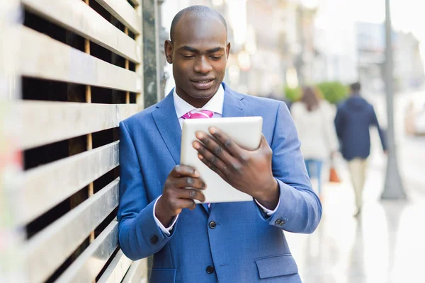 De hombre negro vistiendo traje y corbata mirando su tableta en el fondo urbano — Foto de Stock