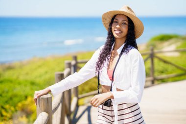 Neşeli yaz kıyafetleri giymiş neşeli bir kadın, güzel plajın yanında neşeli bir şekilde duruyor, geniş bir gülümseme ve güneşli günün sıcaklığının tadını çıkarıyor, arkasında çarpıcı bir okyanus manzarası var.