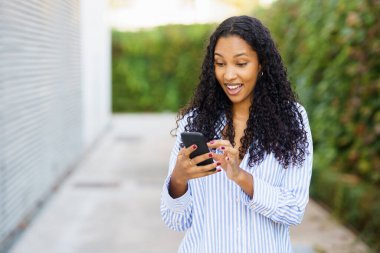 Modern yaşam ve teknolojiyi harmanlayan canlı bir açık havada kıvırcık saçlı neşeli bir kadın akıllı telefonuna gülümsüyor, başkalarıyla iletişim kurarken heyecanını ve mutluluğunu sergiliyor.