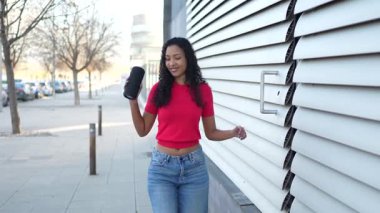 Canlı bir şehir ortamında, kırmızı renkli bluzlu ve mavi kotlu şık genç bir kadın hareketli yaşam tarzında kaygısız bir zevk ve özgürlüğün somutlaştırıcısı, taşınabilir bir hoparlörle neşe içinde dans ediyor.