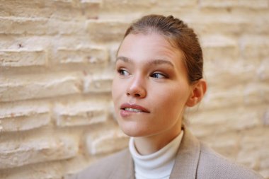 Zarif bir ceket ve klasik balıkçı yaka giyen sakin genç bir kadın zarif bir şekilde dokulu bir duvarın yanında duruyor. Doğuştan gelen zarafetini sergiliyor ve benzersiz bir şekilde şık bir duruş sergiliyor.