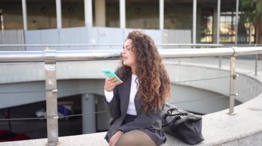 Şık bir genç kadın, modern yaşam tarzı, teknoloji ve çağdaş tarzın mükemmel bir karışımını göstererek, hareketli bir şehir manzarasında akıllı telefonunu kullanırken gelişigüzel oturuyor.