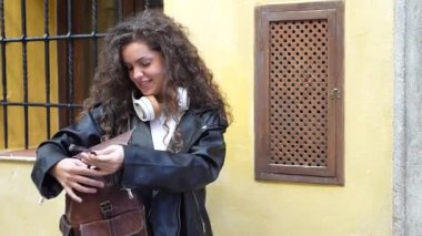 Modaya uygun genç bir kadın parlak siyah deri bir ceket giyer ve parlak sarı bir duvarın önünde şık kahverengi sırt çantasını inceler ve sahneye enerji ve canlı bir atmosfer ekler.