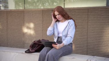 Genç bir kadın tabletini kullanırken gülümseyen güzel bir açık hava ortamında bankta oturuyor. Sıradan giyinerek, sıcak güneş ışığında boş vaktinin tadını çıkarıyor, kendini mutlu ve rahat hissediyor.